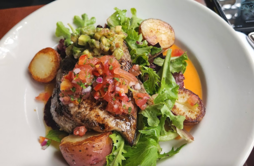 A plate of grilled fish on a bed of greens, topped with salsa and avocado, accompanied by roasted potatoes.