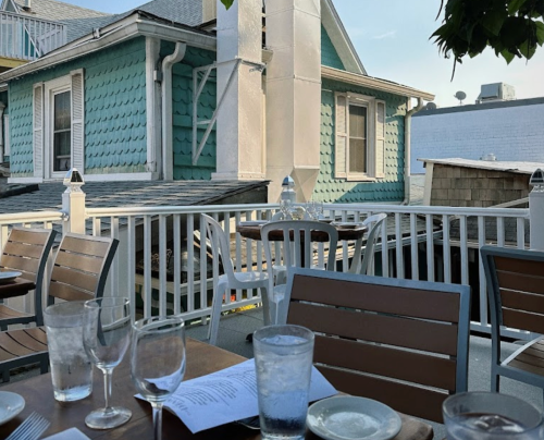 Outdoor dining area with wooden tables, glasses, and a view of a turquoise house and rooftops in the background.