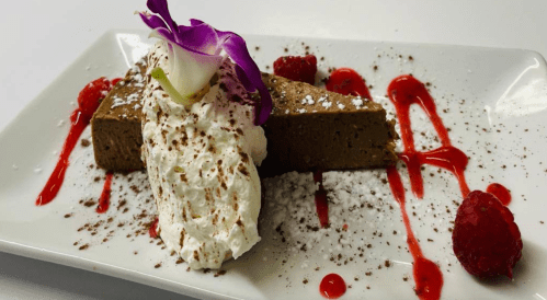 A slice of chocolate mousse cake topped with whipped cream, raspberries, and a purple flower on a white plate.