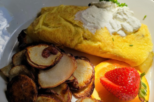 A plate featuring a fluffy omelette topped with sour cream, alongside crispy potatoes, a slice of orange, and a strawberry.