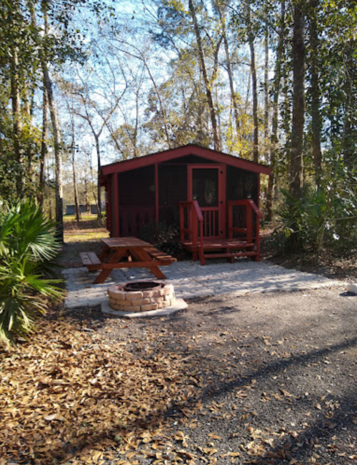 A red cabin surrounded by trees, with a picnic table and fire pit in front on a gravel path.