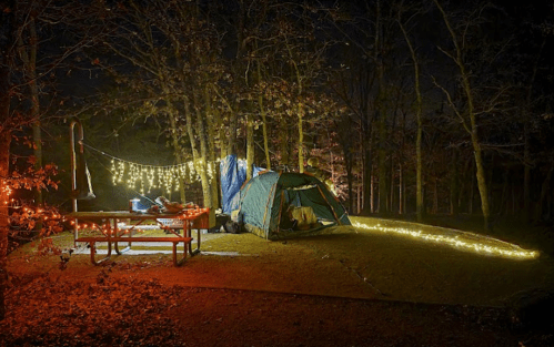 A cozy campsite at night, featuring a tent, picnic table, and string lights illuminating the surrounding trees.