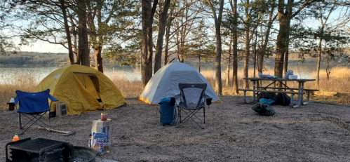 Two tents and camping chairs by a lake, surrounded by trees, with a picnic table and fire pit nearby.