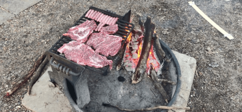 Grilling raw meat and sausages over an open fire in a campfire pit.