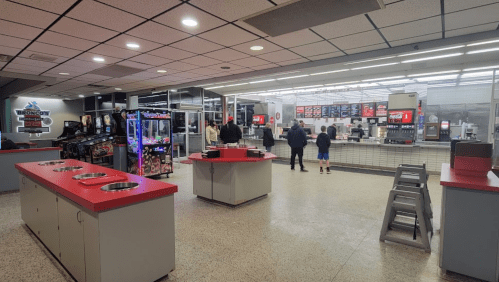 A modern concession stand with a counter, arcade games, and customers waiting in line. Brightly lit and spacious interior.