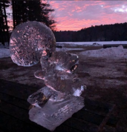 An ice sculpture of a squirrel against a sunset backdrop, with trees and snow in the foreground.