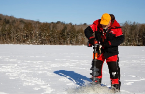 A person in winter gear drills a hole in a snowy landscape using an ice auger. Trees are visible in the background.