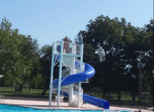A blue water slide at a pool with a person standing at the top, surrounded by trees and clear blue sky.
