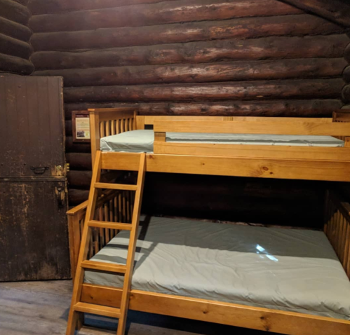 A rustic cabin interior featuring a wooden bunk bed with empty mattresses and a dark wooden door in the background.