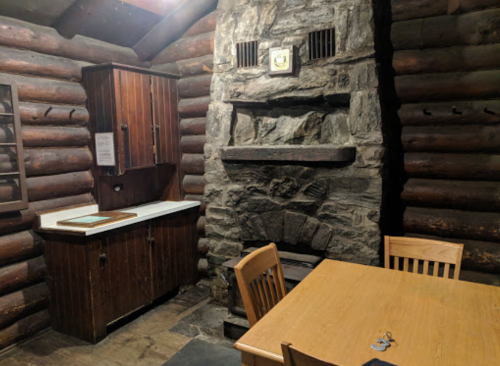 Cozy log cabin interior featuring a stone fireplace, wooden furniture, and a rustic kitchen area.