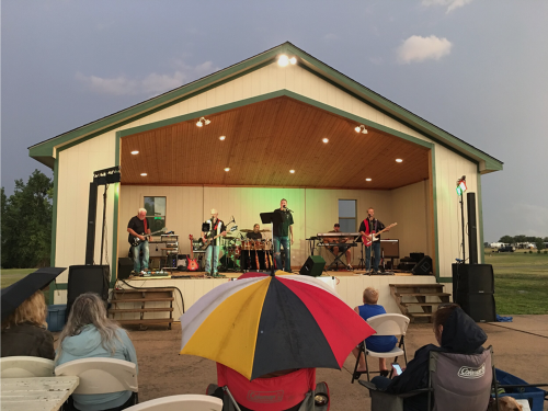 A band performs on stage under a cloudy sky, while an audience sits with umbrellas in front of them.