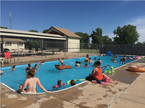 A busy outdoor pool scene with children and adults swimming and relaxing on pool floats under a clear blue sky.