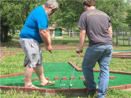 Two men playing mini-golf on a green course, focusing on their shots with putters in hand.