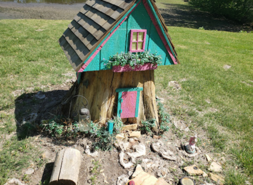 A whimsical, colorful miniature house with a sloped roof, surrounded by greenery and decorative stones.