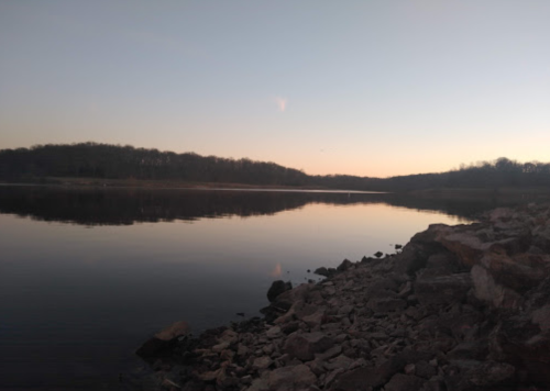 A calm lake at sunset, with rocky shorelines and trees silhouetted against the horizon.