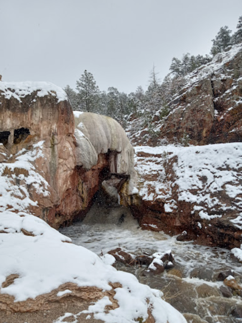 A snowy landscape featuring a frozen waterfall cascading over rocky terrain, surrounded by pine trees.