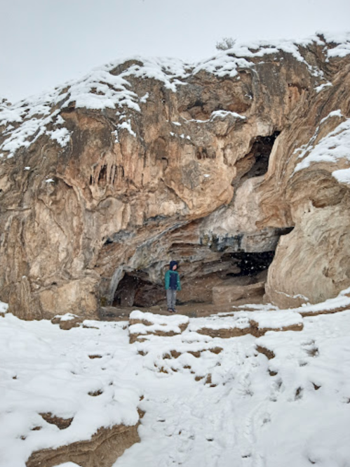 A person stands in front of a snow-covered cave entrance on a rocky hillside.