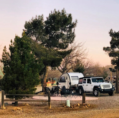 A camper and Jeep parked near trees, with outdoor seating in a serene, gravel area at sunset.