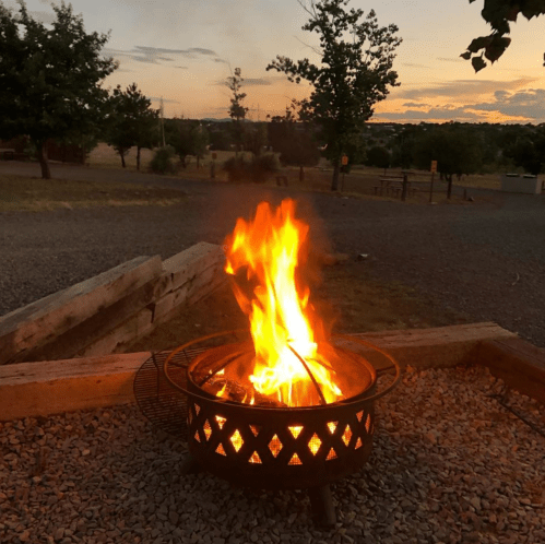 A vibrant campfire burns in a metal fire pit, surrounded by gravel and trees, with a sunset in the background.