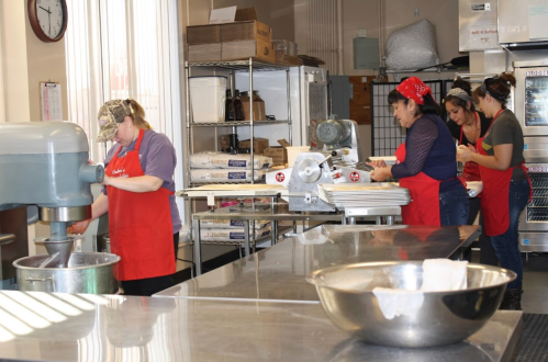 A busy kitchen with four women in red aprons working with a mixer, slicer, and baking trays.