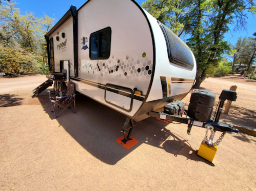 A parked travel trailer with a small outdoor seating area, surrounded by trees and gravel.