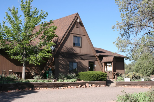 A brown, A-frame house surrounded by trees and shrubs, with a gravel path leading to the entrance.