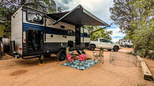 A camper trailer with an awning set up in a campsite, featuring chairs and a truck parked nearby.