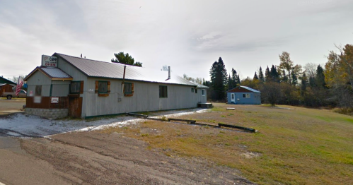A gray building with a metal roof and a blue house nearby, set in a grassy area with trees in the background.