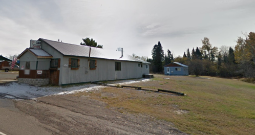 A weathered building with a metal roof beside a gravel road, surrounded by trees and a small blue house in the background.