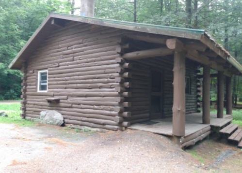 A rustic log cabin surrounded by trees, featuring a porch and a gravel path.