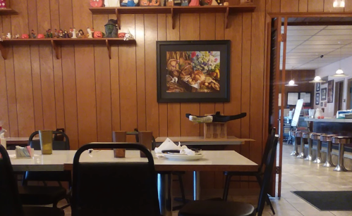 Cozy restaurant interior with wooden walls, a framed painting, and empty tables set for dining.