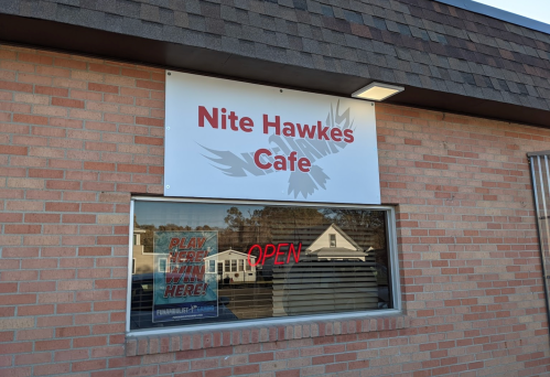 Sign for "Nite Hawkes Cafe" on a brick building, with an illuminated "OPEN" sign in the window.