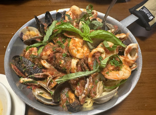 A seafood pasta dish with shrimp, mussels, clams, and herbs in a rich tomato sauce, served in a metal bowl.