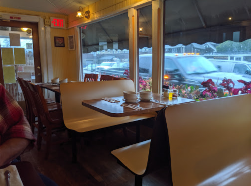 A cozy diner interior with empty tables, plates set, and a view of a rainy street through large windows.