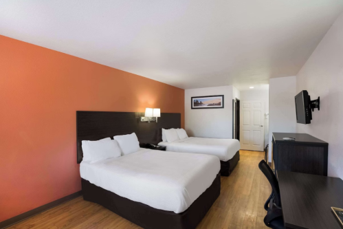 A hotel room with two beds, a desk, and a TV, featuring orange and white walls and wooden flooring.