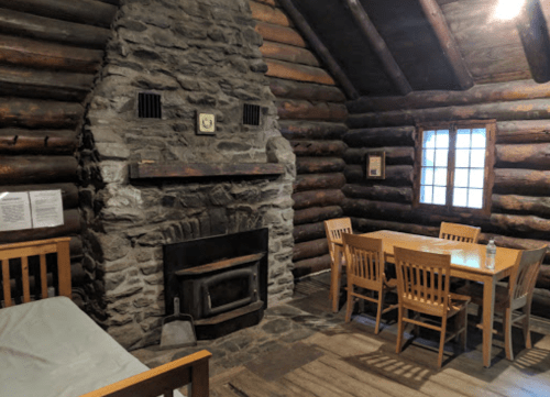 Cozy log cabin interior featuring a stone fireplace, wooden furniture, and a window with natural light.