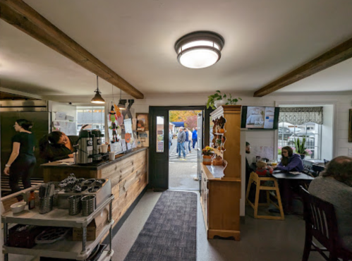 A cozy café interior with a wooden counter, patrons seated, and an open door leading to an outdoor area.