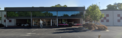 A building with large windows showcasing classic cars, surrounded by trees and a parking lot.