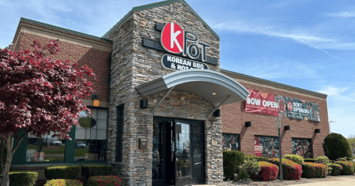 Exterior of K-Pot Korean BBQ & Hot Pot restaurant, featuring a stone facade and banners announcing its opening.