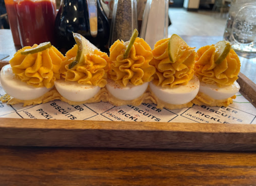 A wooden platter with five dollops of yellow frosting on white biscuits, garnished with green slices and spices.