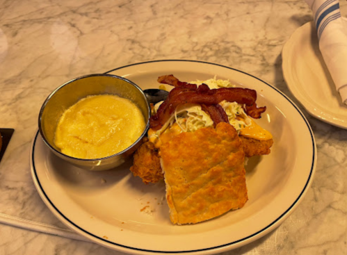 A plate with fried chicken sandwich topped with bacon and coleslaw, served with a side of creamy grits.