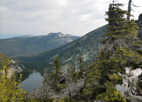 A scenic mountain landscape with lush trees, a lake, and distant peaks under a cloudy sky.
