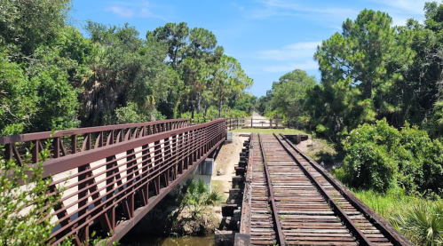 A metal bridge over a creek, with old wooden train tracks beside it, surrounded by lush greenery and blue skies.