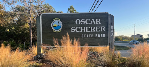 Sign for Oscar Scherer State Park in Florida, featuring a logo and surrounded by tall grass and trees.