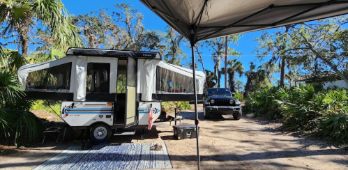 A pop-up camper parked in a sunny campsite surrounded by palm trees and greenery, with a vehicle nearby.