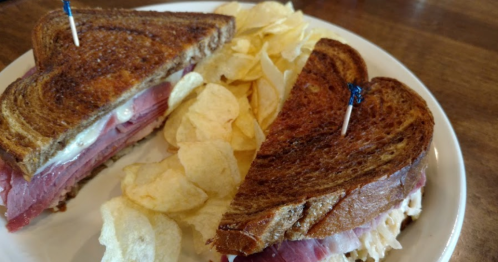 A close-up of a toasted sandwich with layers of meat and cheese, served with a side of potato chips.