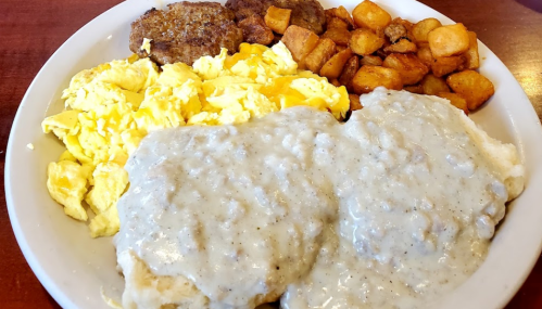 A hearty breakfast plate with scrambled eggs, sausage patties, seasoned potatoes, and biscuits topped with gravy.