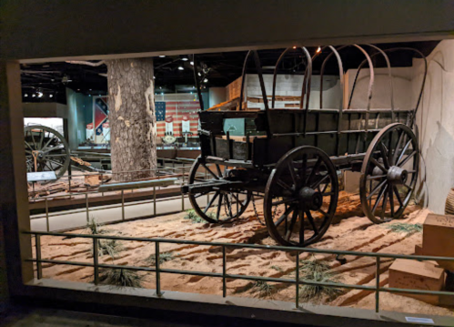 A historic wagon displayed in a museum, surrounded by sand and featuring a backdrop of flags and artifacts.