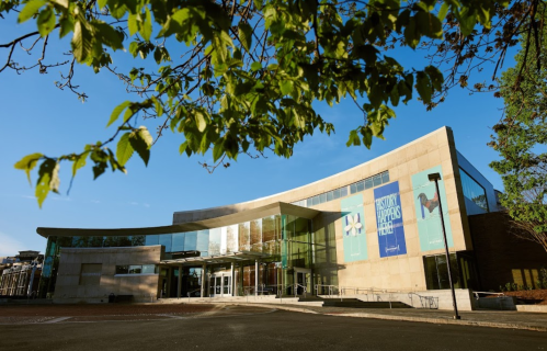 Modern building with large glass entrance, surrounded by trees and blue sky, featuring a colorful banner.