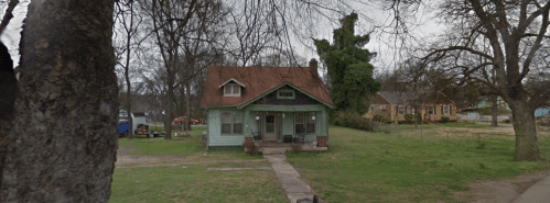 A small, green house with a red roof, surrounded by trees and a grassy yard, on a quiet street.
